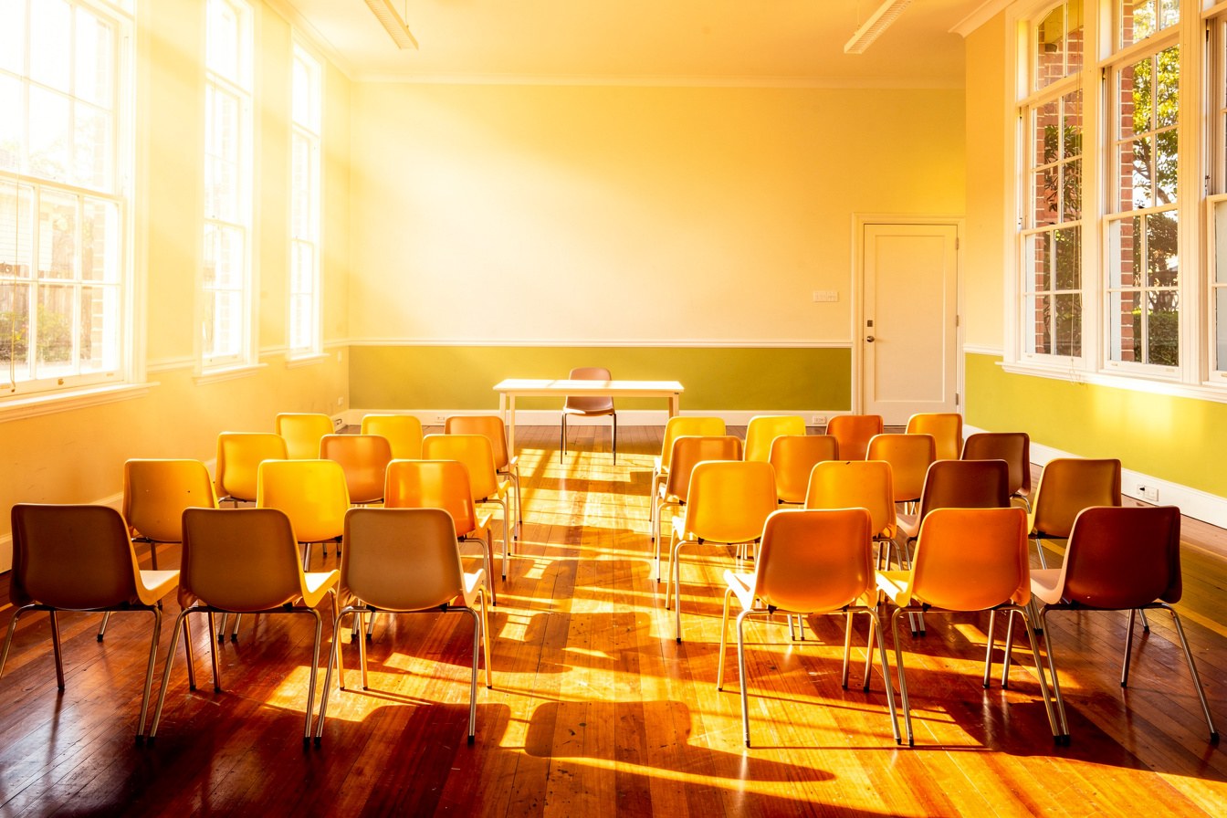 An empty community hall set up with rows of chairs for a Saturday language class, sunlight streaming through tall windows.