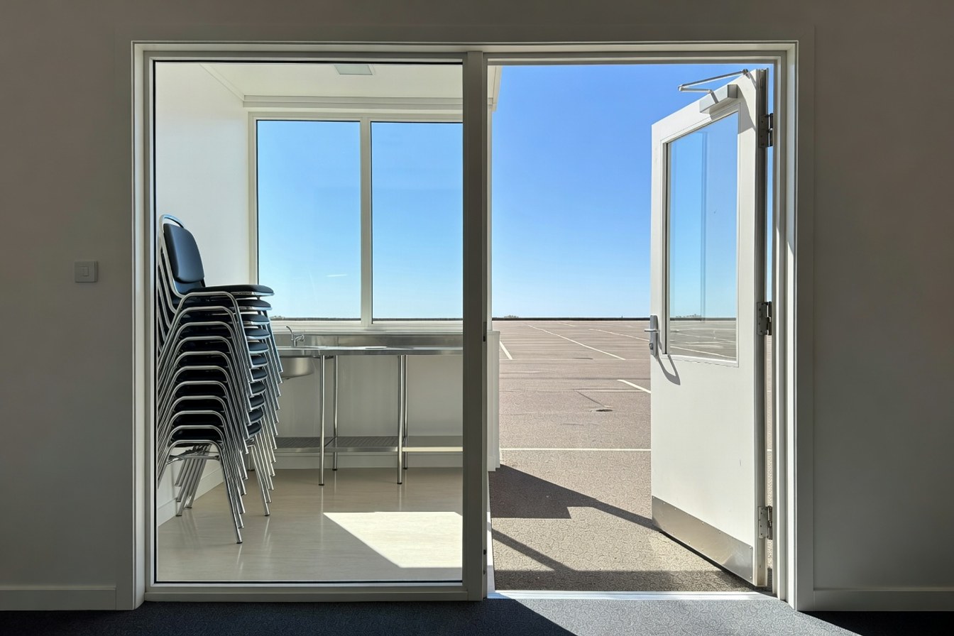An open door of a community hall lets in daylight, one side already closing, suggesting a deadline passing.